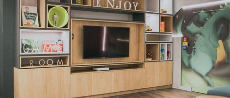 Un área de entretenimiento moderna con un televisor de pantalla plana grande, estanterías de madera con varios objetos y un mural en la pared.