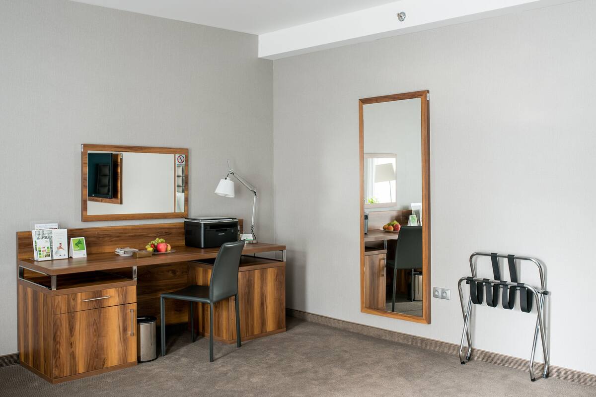 A hotel room with a wooden desk, a chair, a mirror, a lamp, and a luggage rack.
