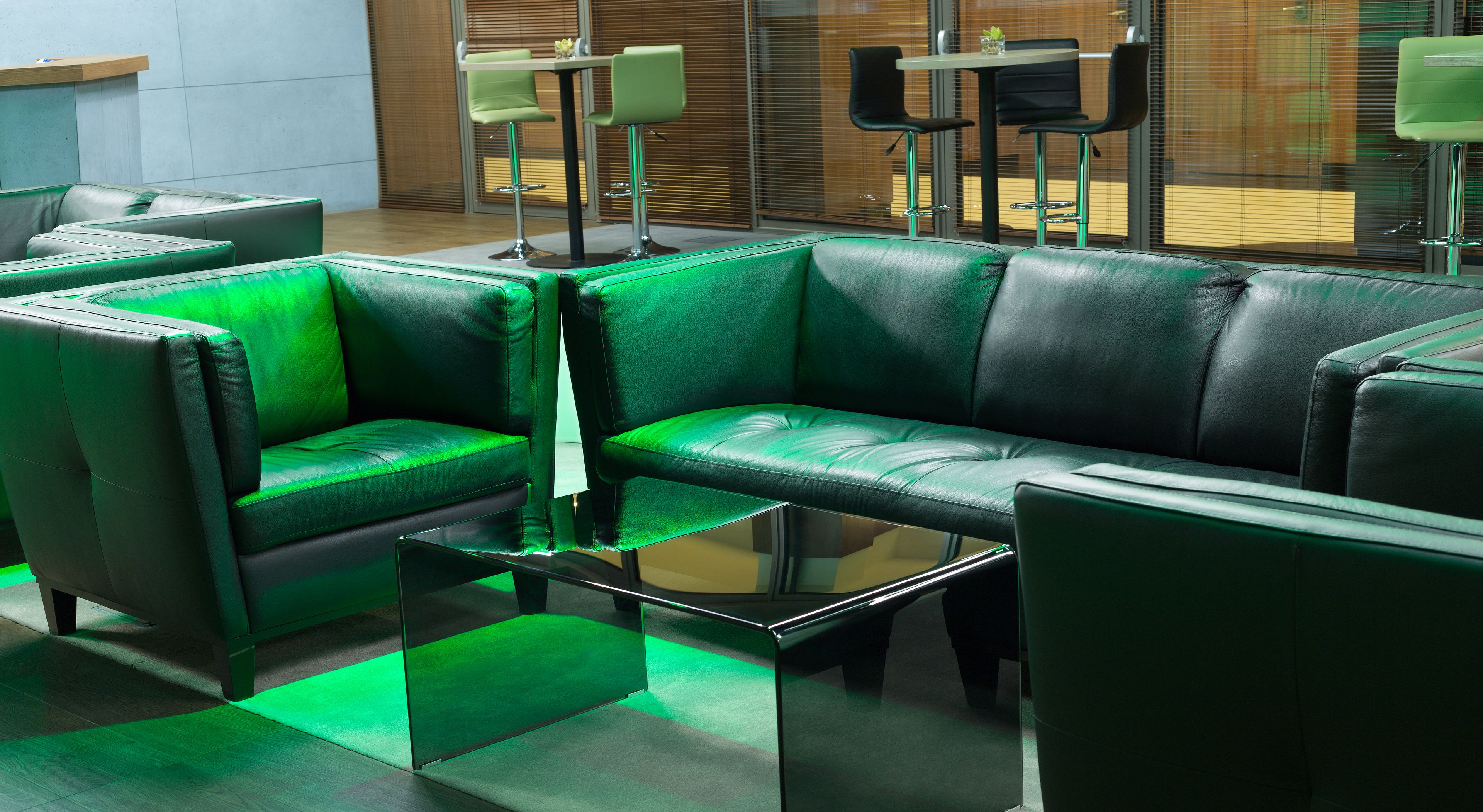 A modern lounge area with green leather sofas and chairs, a glass coffee table, and a wooden floor.