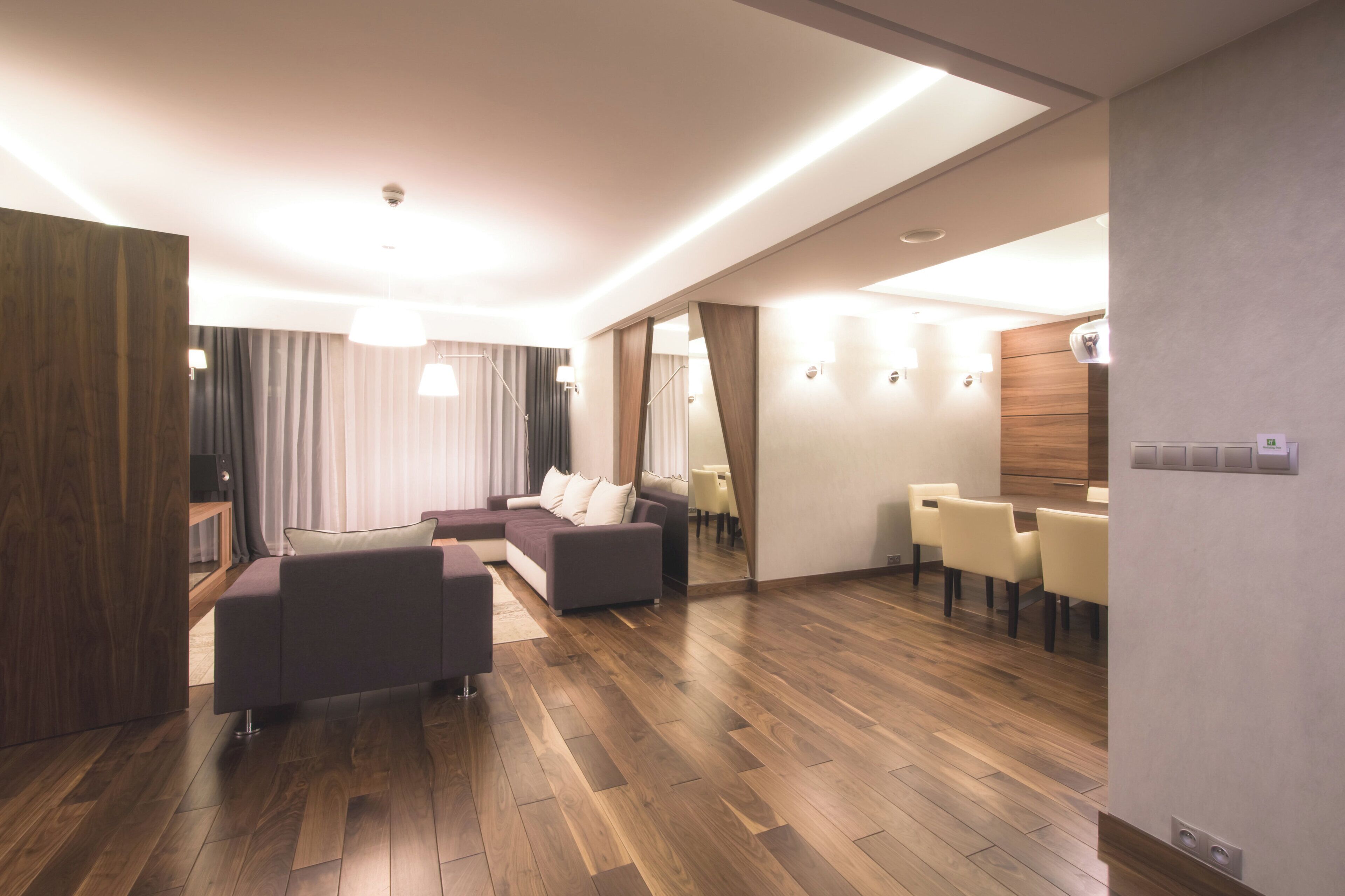 A modern hotel room with a wooden floor, a sofa set, a dining area with chairs, and a large mirror.