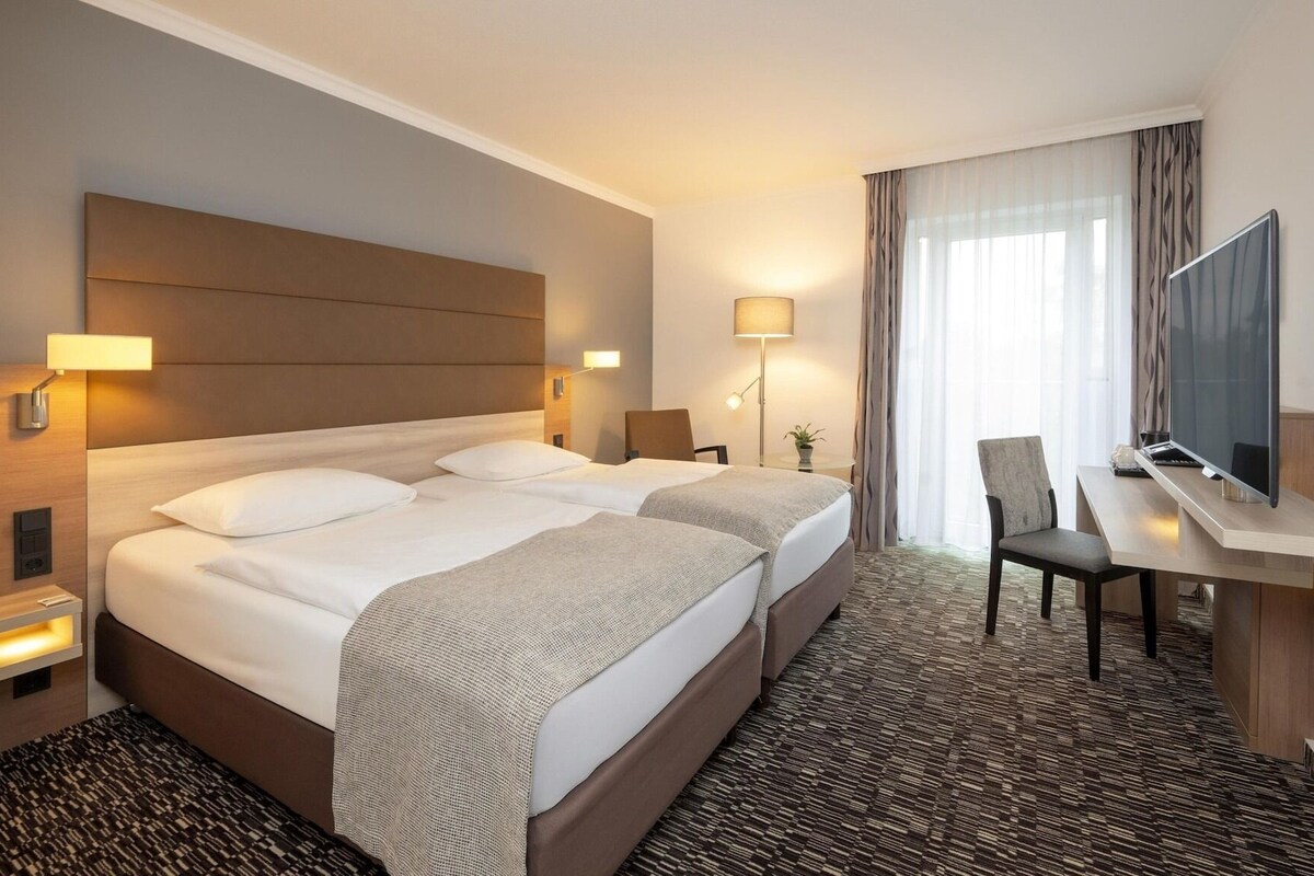 Ein Hotelzimmer mit einem großen Bett, Nachttischen, einem Schreibtisch, einem Stuhl, einem Fernseher und einem Fenster mit Vorhängen.