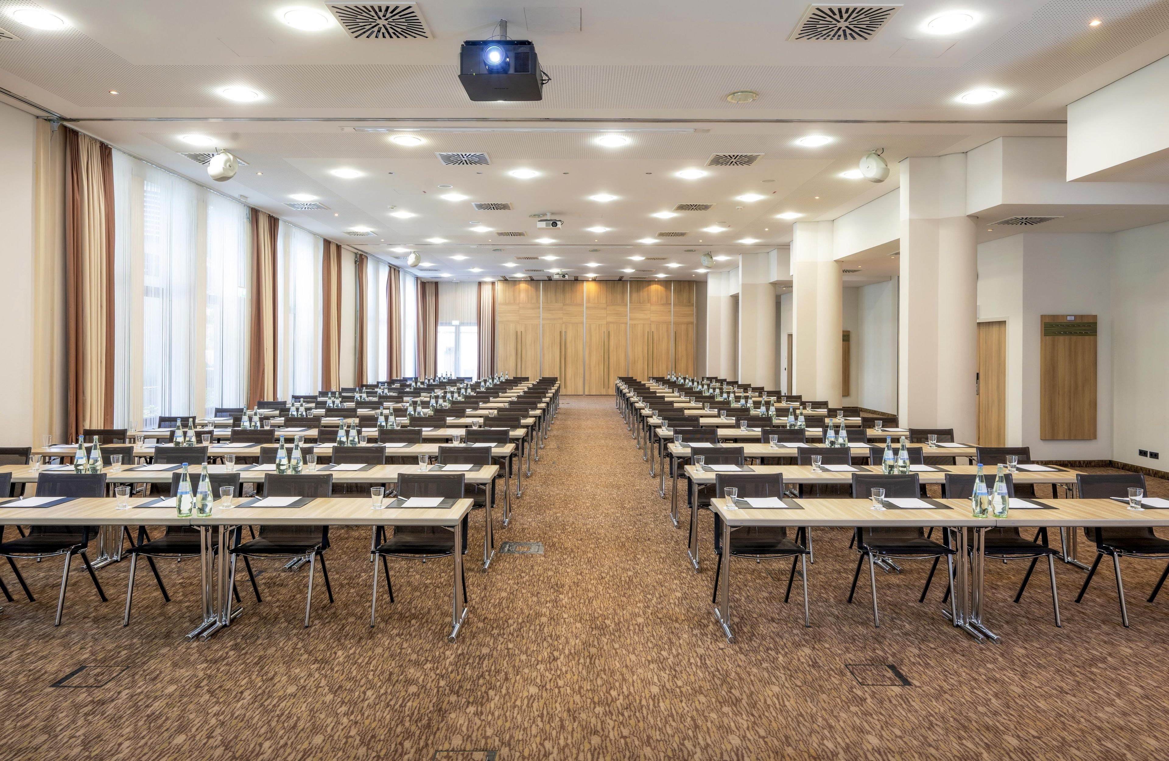 Ein Konferenzraum mit Tischen und Stühlen, einem Projektor und großen Fenstern mit Vorhängen.