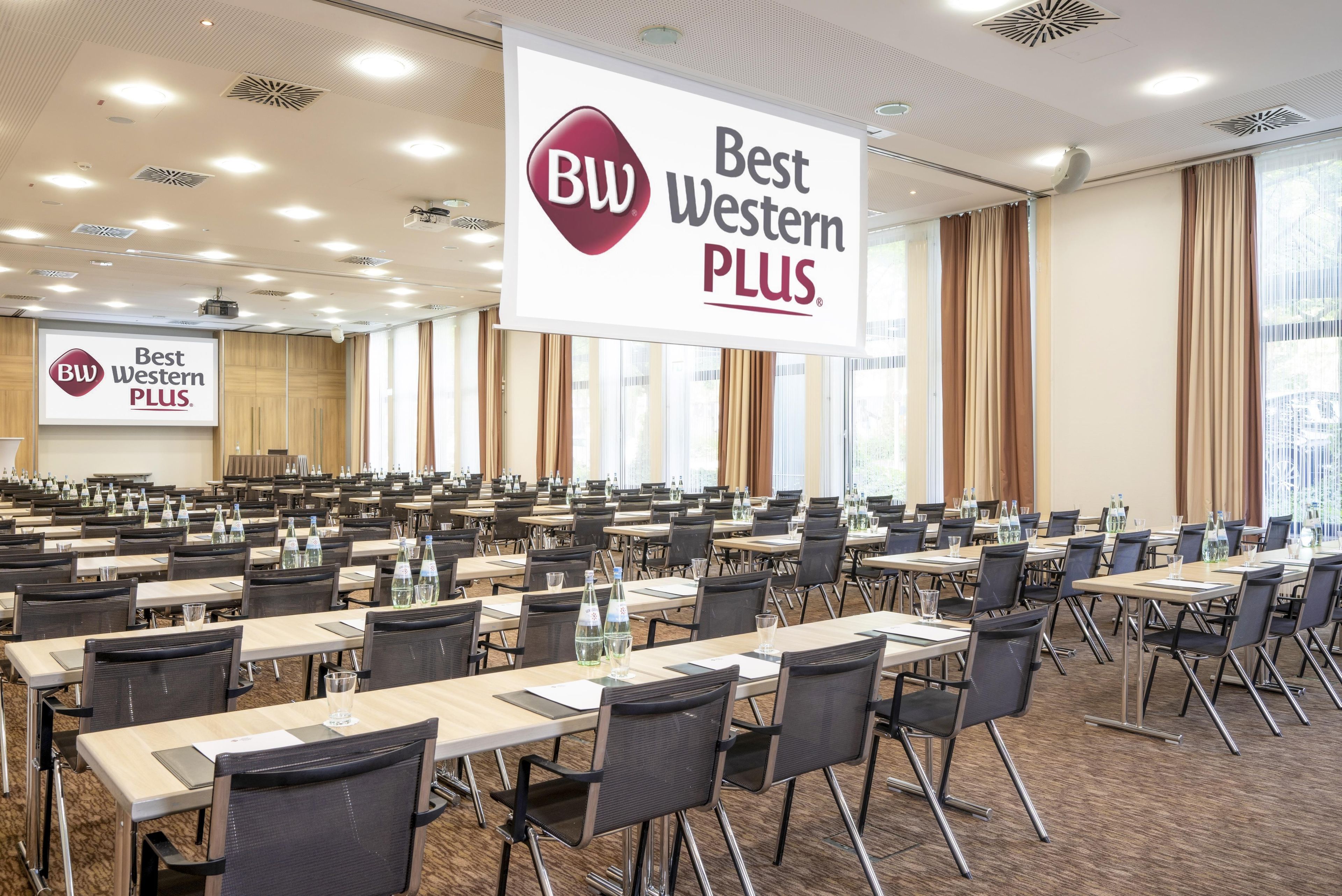 Ein Konferenzraum mit Reihen von Tischen und Stühlen, einem großen Banner mit der 'Best Western PLUS'-Marke und einer Projektionsleinwand im Hintergrund.