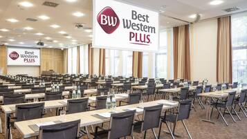 Ein Konferenzraum mit Reihen von Tischen und Stühlen, einem großen Banner mit der 'Best Western PLUS'-Marke und einer Projektionsleinwand im Hintergrund.