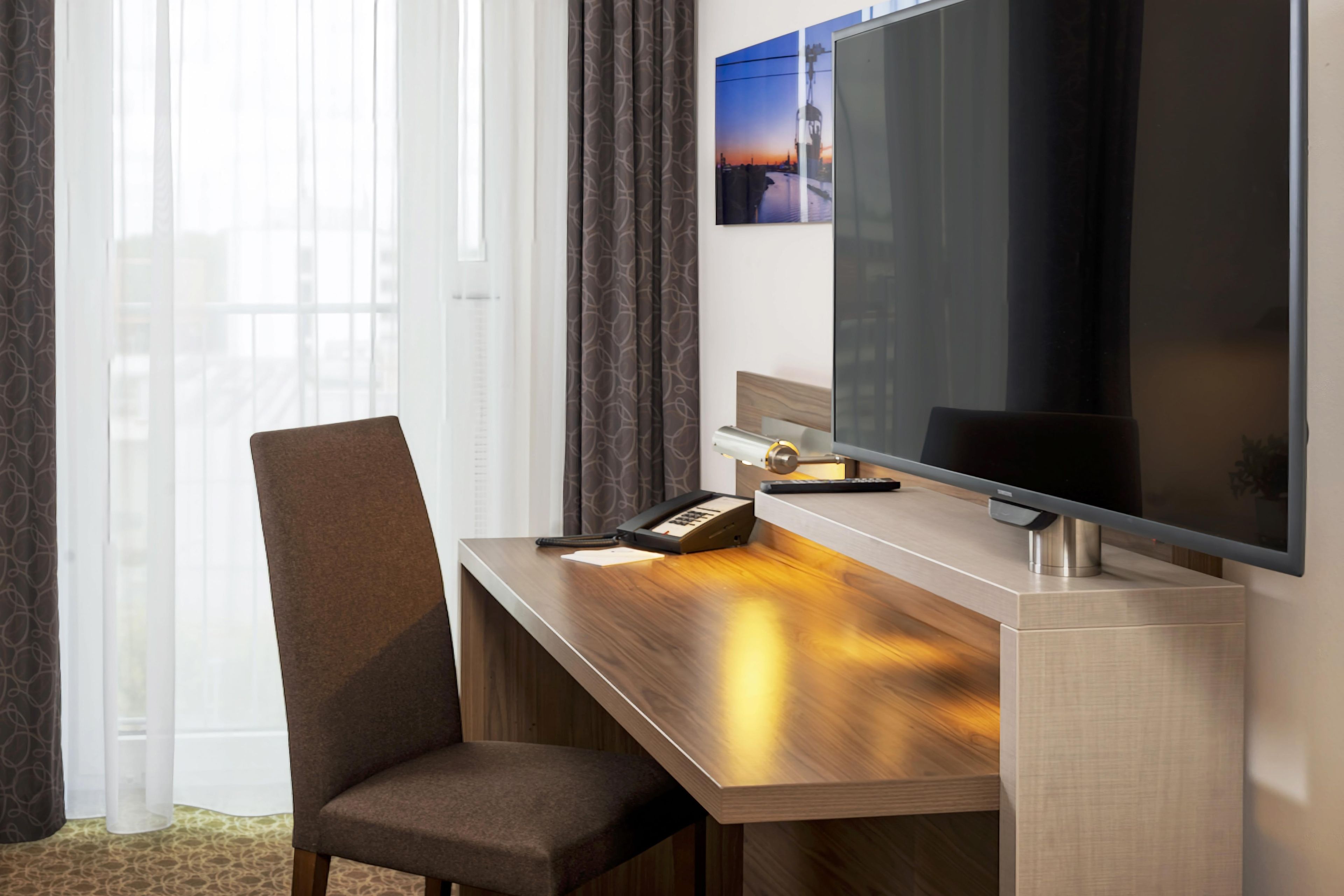Habitación de hotel con un escritorio de madera, una silla, un televisor de pantalla plana y una ventana con cortinas.