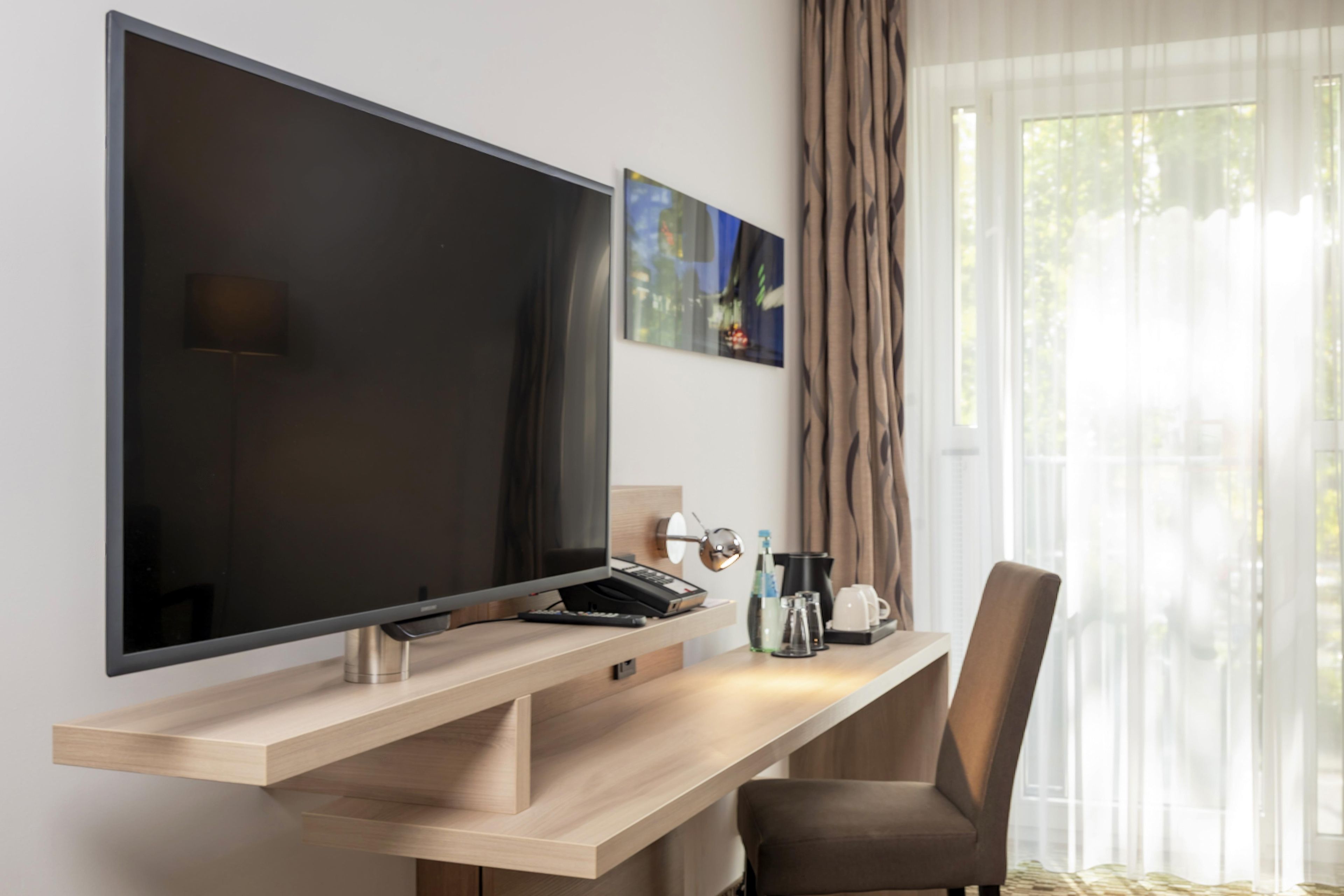 Ein modernes Hotelzimmer mit einem großen Flachbildfernseher an einem Holztisch, einer Kaffeemaschine, einer Fernbedienung und einem Sessel.