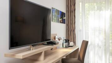 Ein modernes Hotelzimmer mit einem großen Flachbildfernseher an einem Holztisch, einer Kaffeemaschine, einer Fernbedienung und einem Sessel.