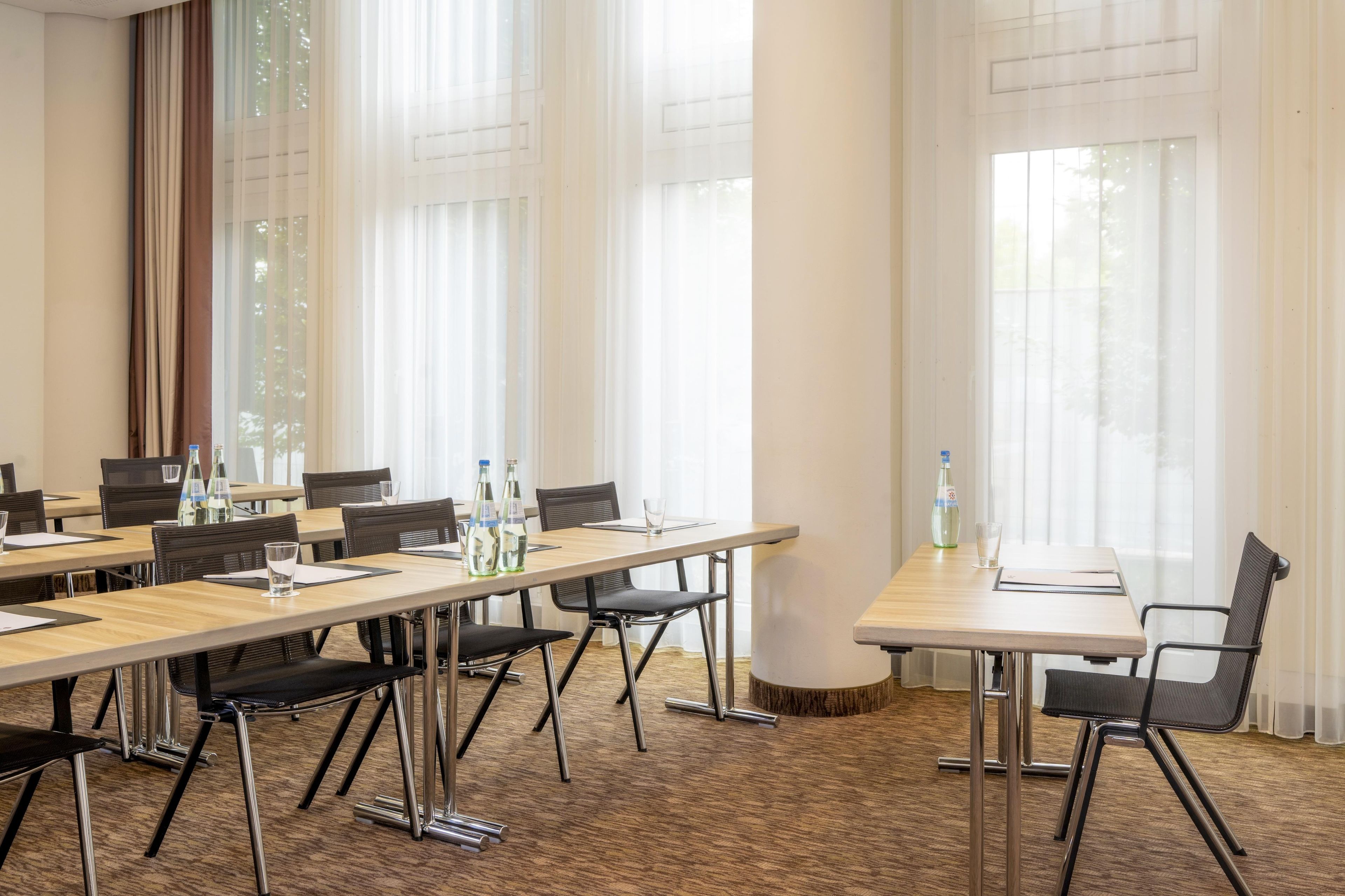 A conference room with multiple tables and chairs, large windows with sheer curtains, and a carpeted floor.