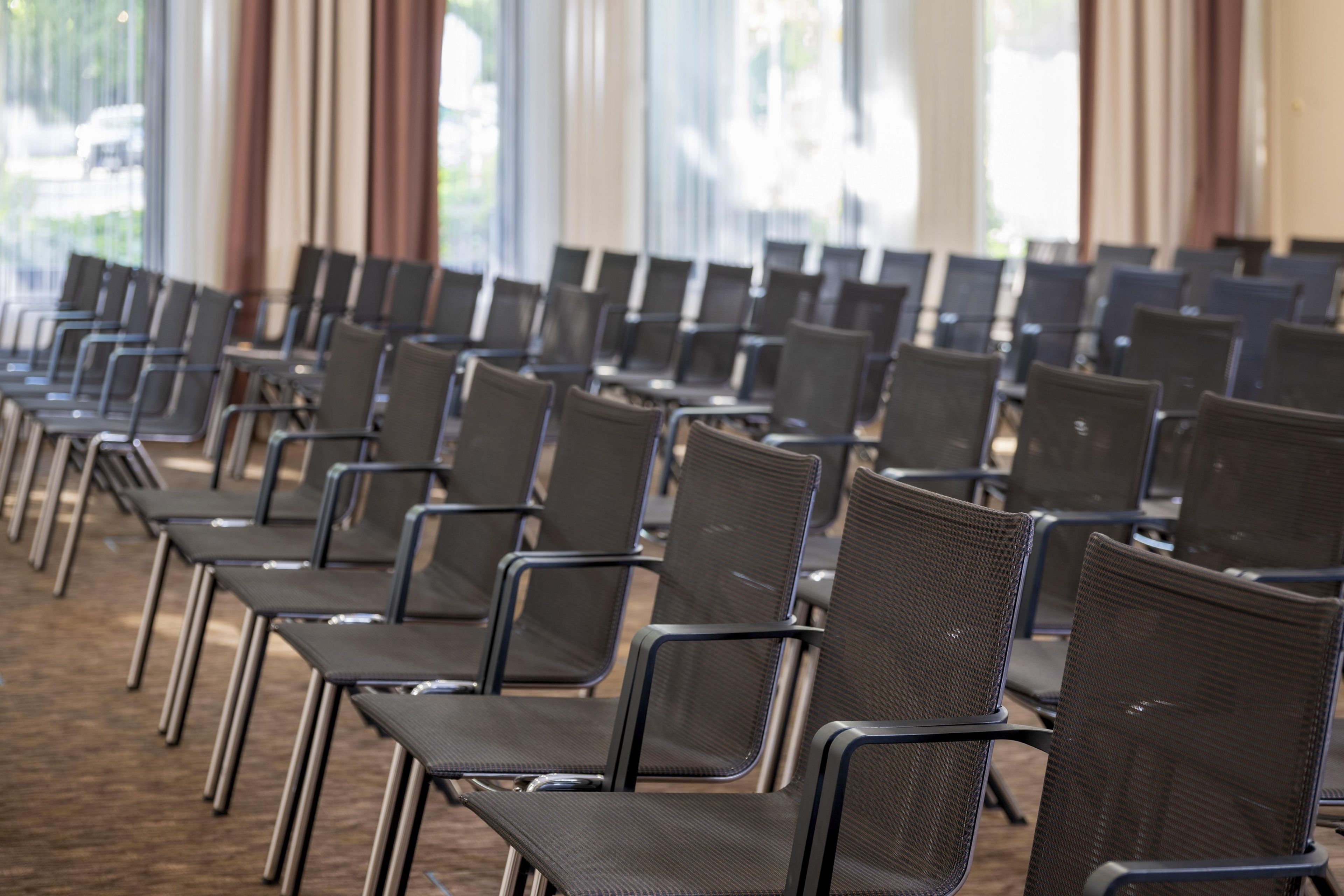 Reihenweise Stühle in einem Konferenzraum mit großen Fenstern und Vorhängen.