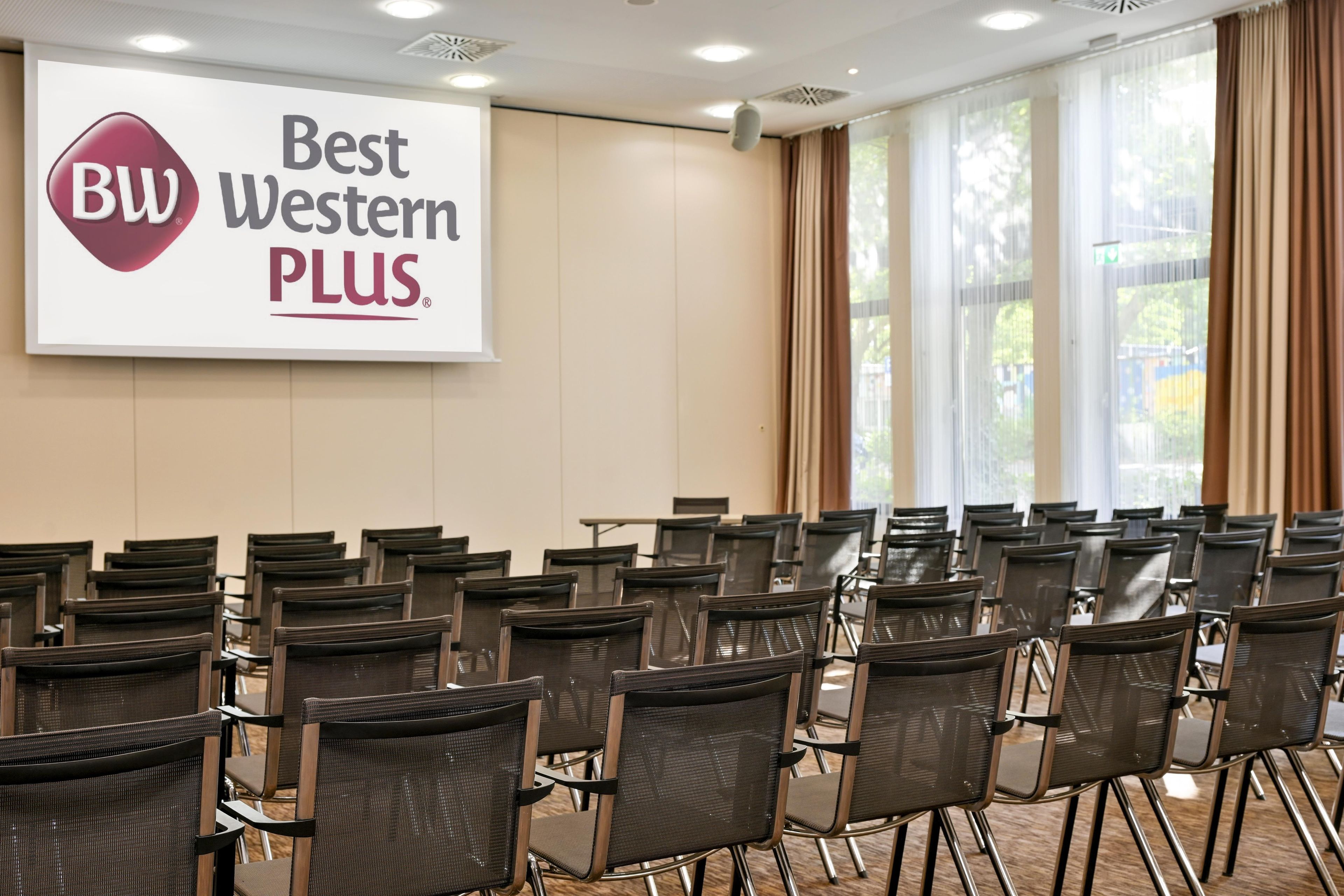 Ein Konferenzraum mit Reihen von Stühlen, einem großen Bildschirm, der das Logo von Best Western PLUS zeigt, und einem Fenster mit Vorhängen.