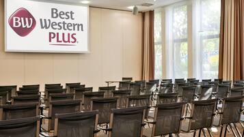 Ein Konferenzraum mit Reihen von Stühlen, einem großen Bildschirm, der das Logo von Best Western PLUS zeigt, und einem Fenster mit Vorhängen.