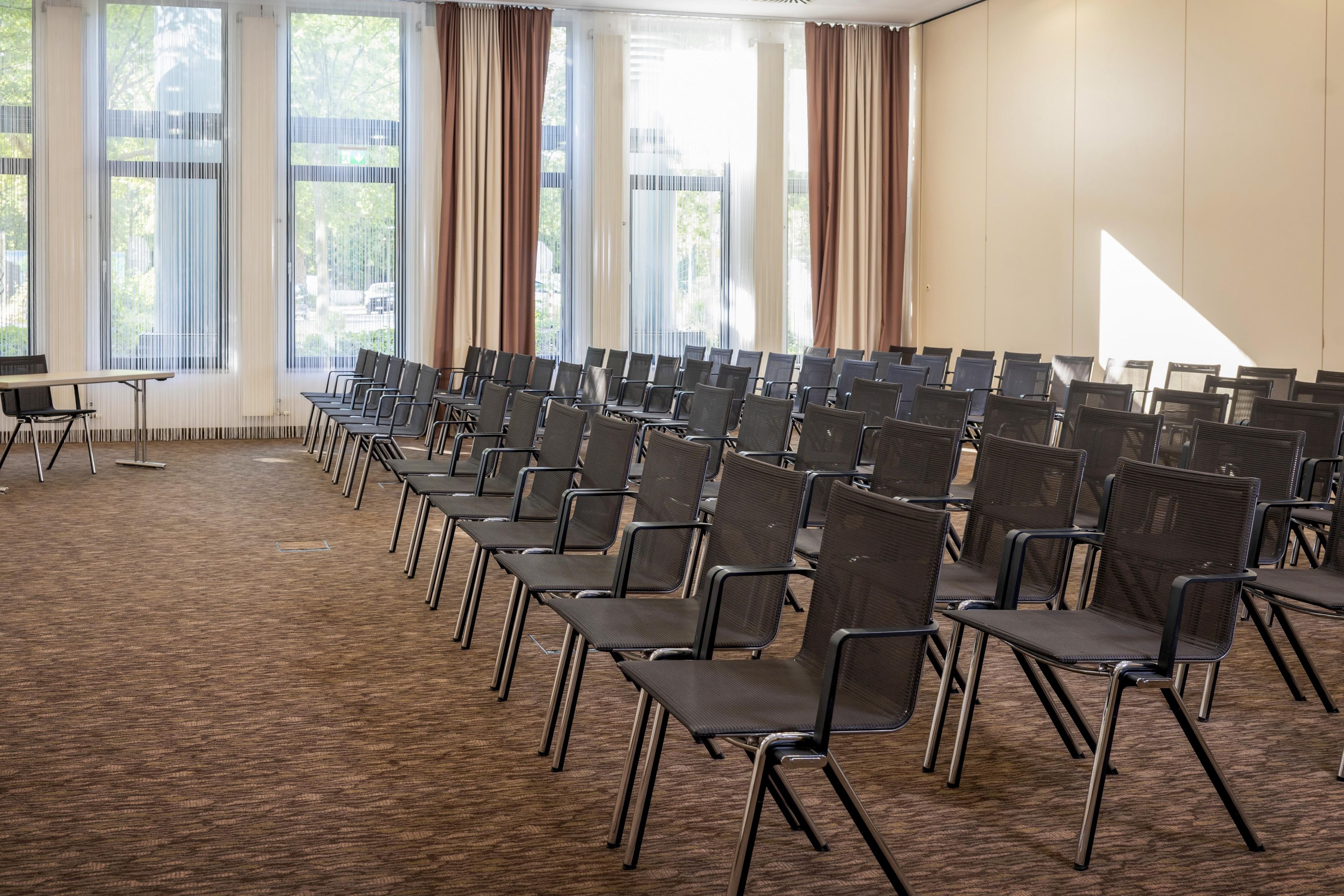 Ein Konferenzraum mit Stuhlreihen, großen Fenstern mit Vorhängen und einem Teppichboden.