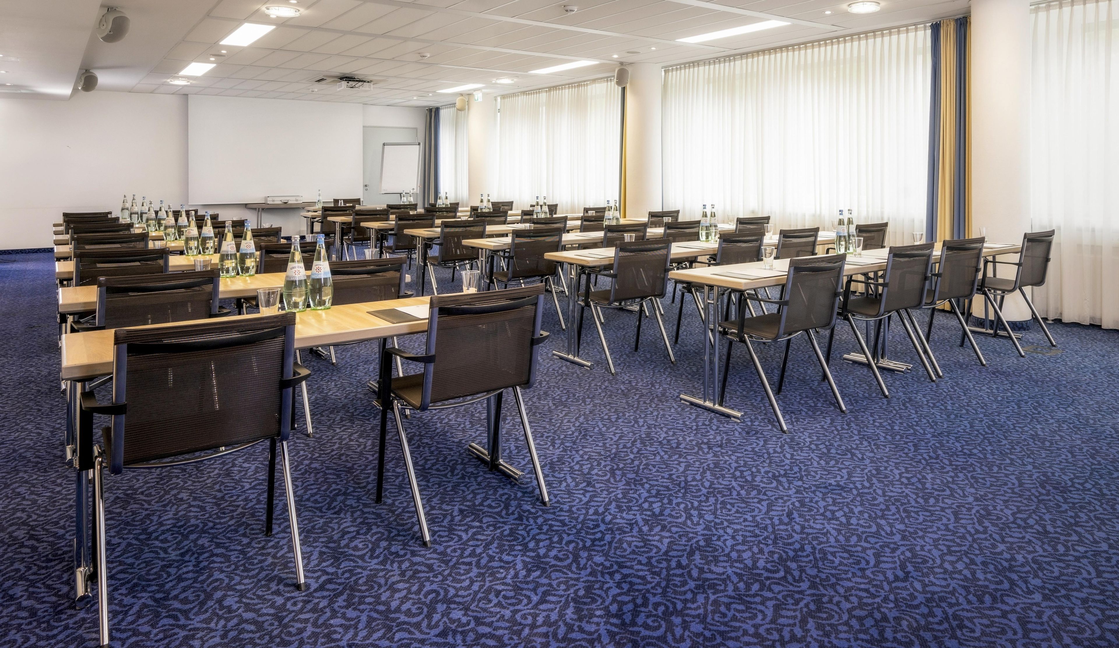 Une salle de conférence équipée de tables et de chaises, d’un écran de projection et d’un tableau blanc.