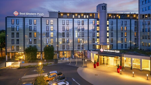 Ein modernes Hotel mit mehreren Etagen, einem markanten Eingang und einem Parkplatz.