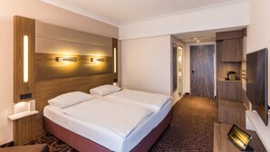 A hotel room with a double bed, wooden headboard, bedside tables, a flat-screen TV, and a small desk with a lamp. - Best Western Plus Hotel Köln City (Cologne)