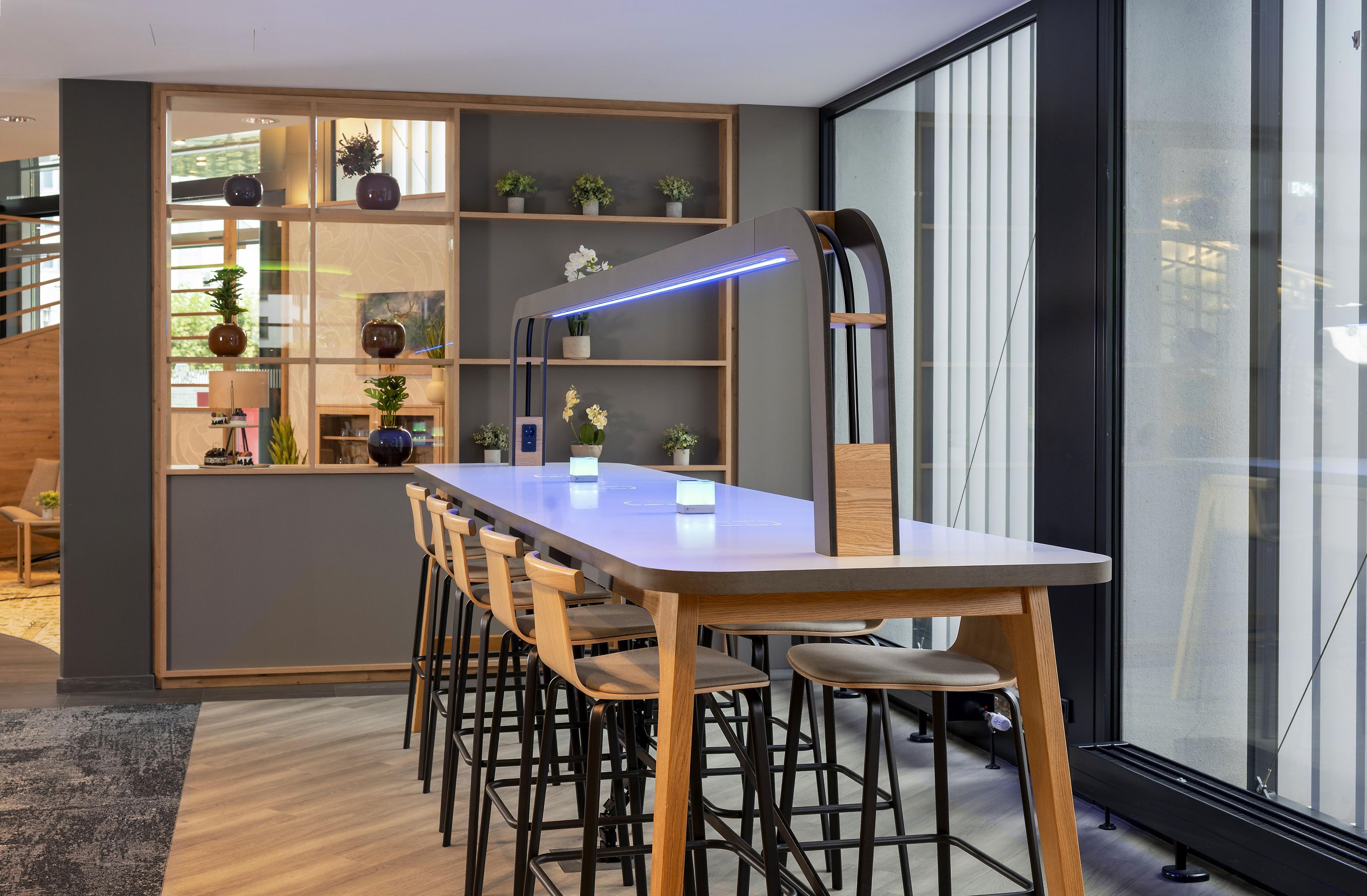Un interior moderno con una mesa larga, múltiples sillas, un escritorio con computadora y estantes con plantas.