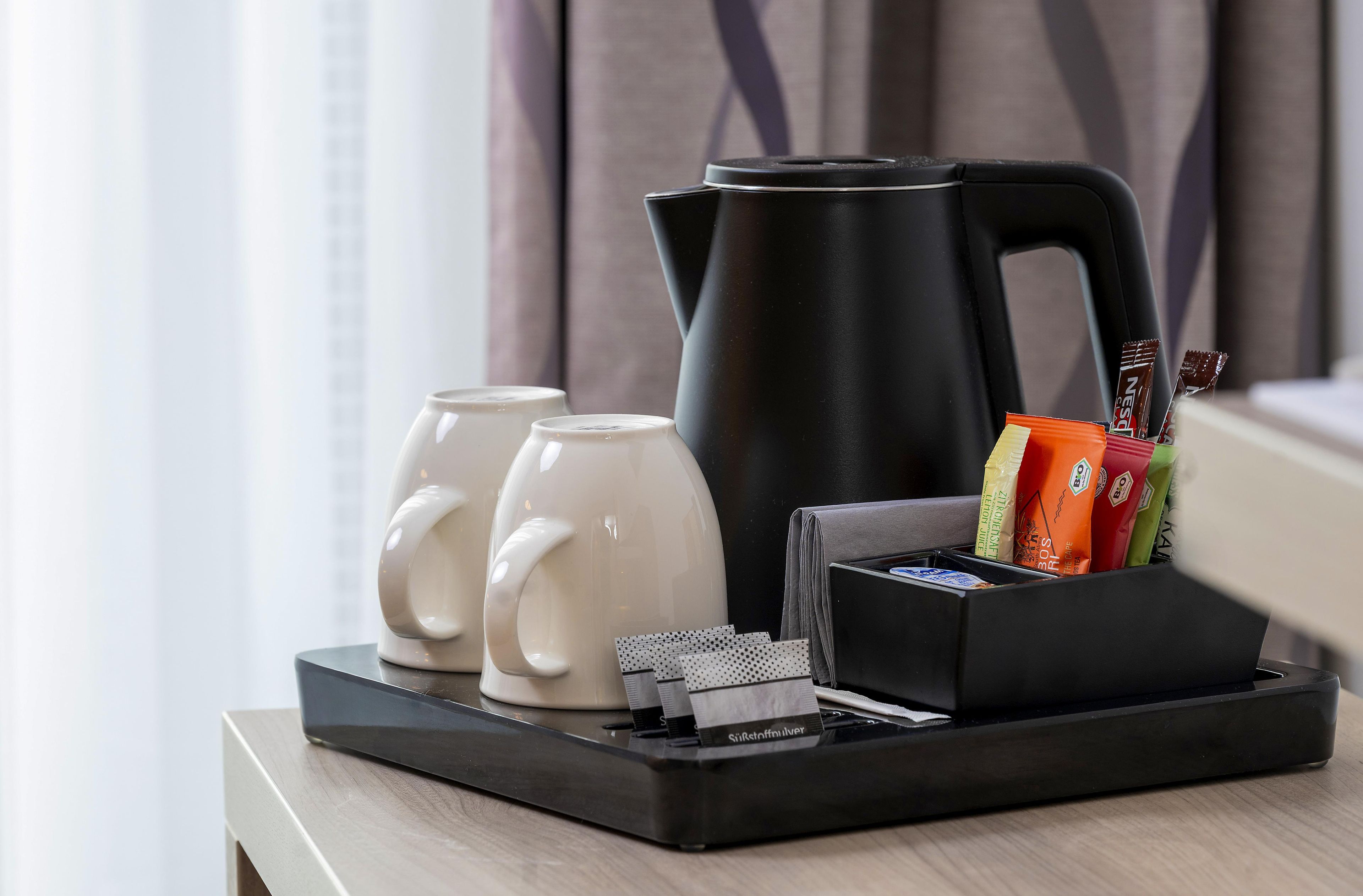 Una bandeja con una tetera eléctrica negra, dos tazas blancas y una caja que contiene varios objetos, incluyendo una caja de pañuelos y algunos bocadillos.