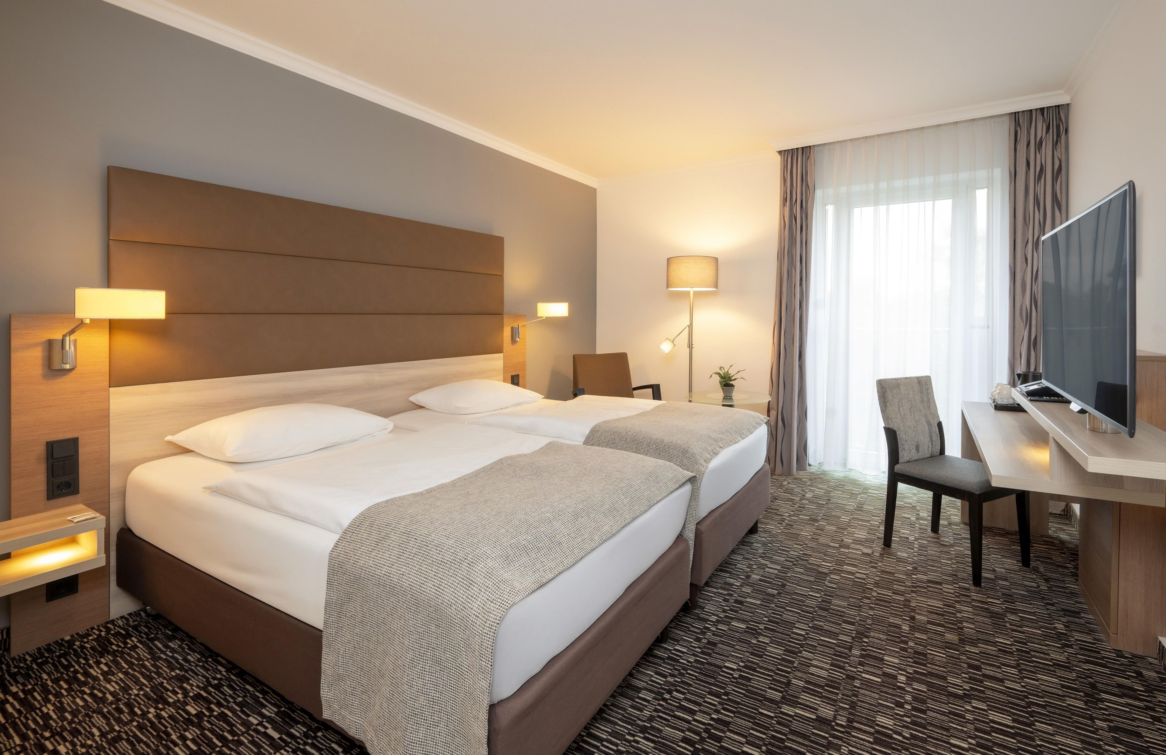 Ein Hotelzimmer mit einem großen Bett, Nachttischen, einem Schreibtisch mit Stuhl, einem Fernseher und einem Fenster mit Vorhängen.