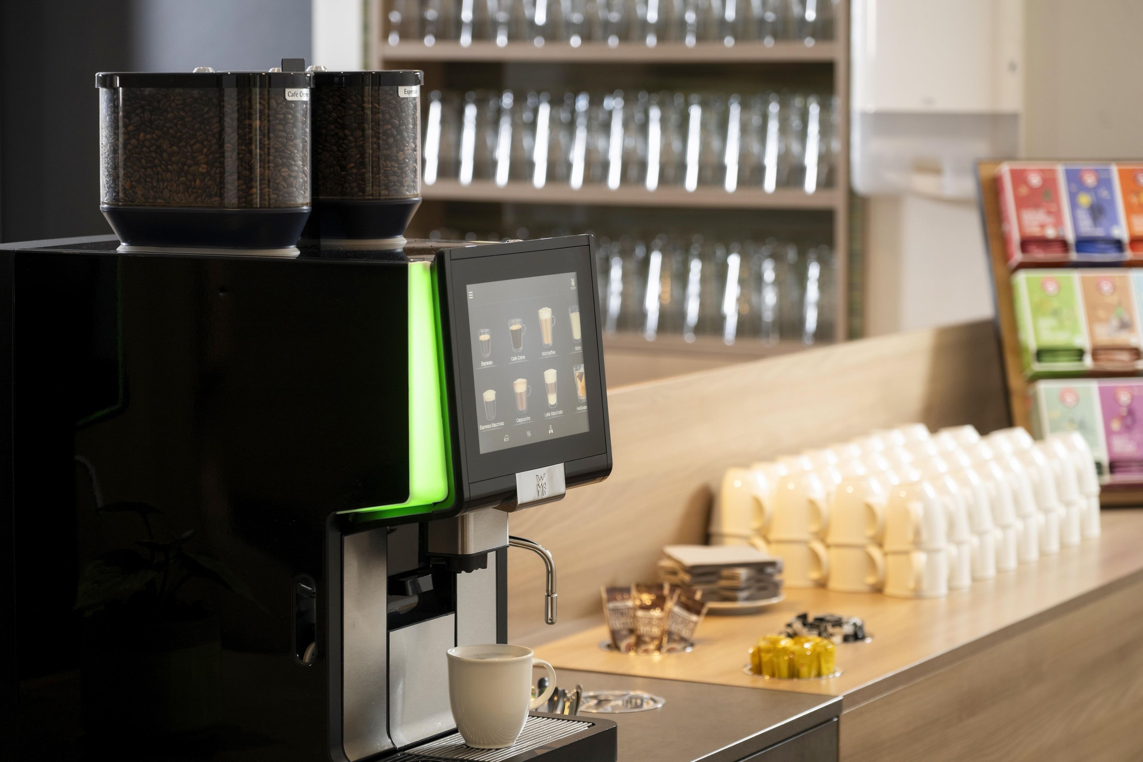 Una cafetera con pantalla digital, una taza de café y una fila de vasos desechables sobre una encimera.