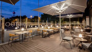 Ein Terrassenbereich im Freien mit Tischen, Stühlen, Sonnenschirmen und Blick auf die Skyline der Stadt bei Einbruch der Dunkelheit.