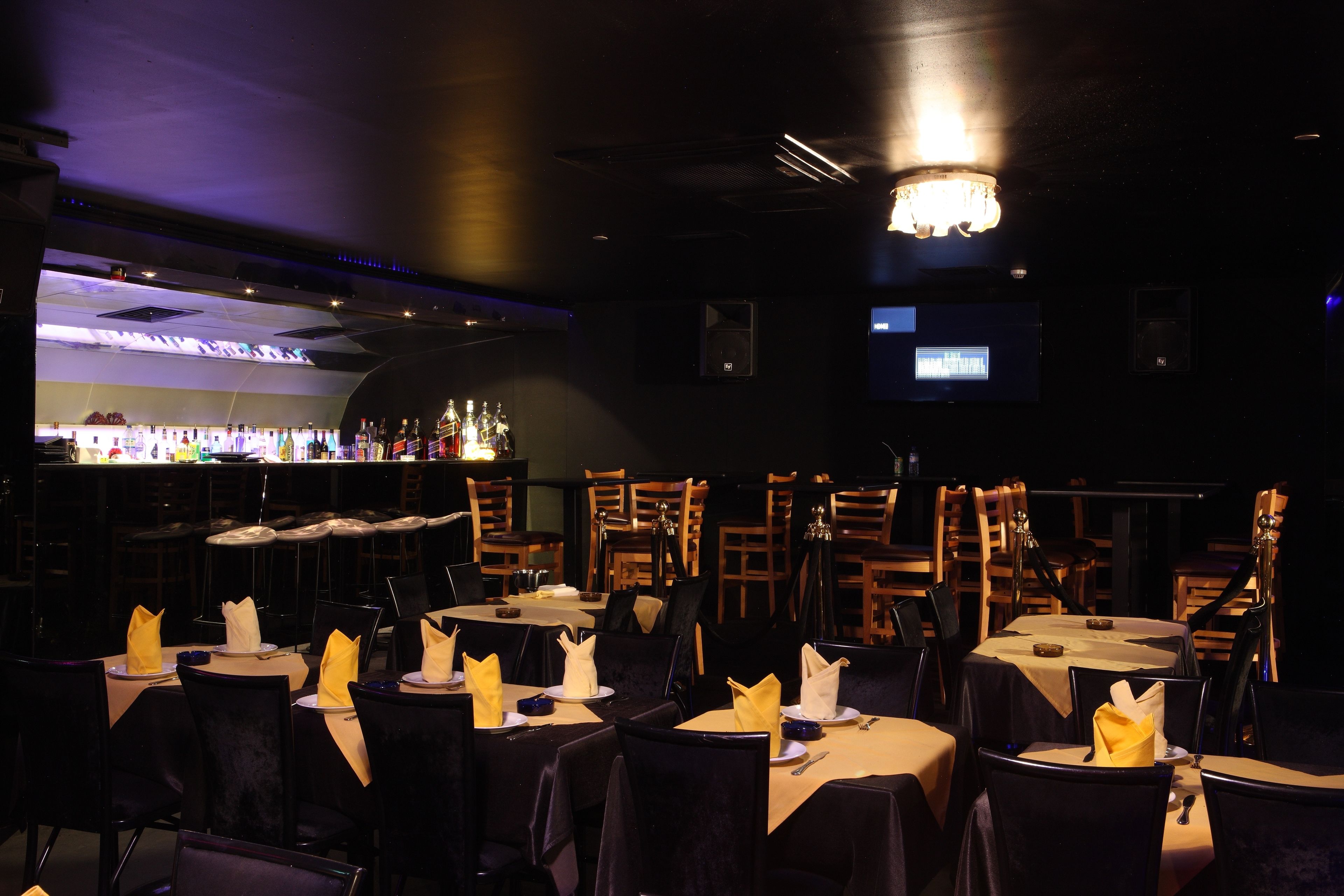 A dimly lit restaurant with tables set for dining, a bar area in the background, and a television screen on the wall.