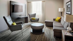 A modern hotel room with a flat-screen TV mounted on the wall, a grey armchair with a yellow pillow, a round ottoman, a grey sofa with yellow pillows, and a side table with a lamp and remote control.