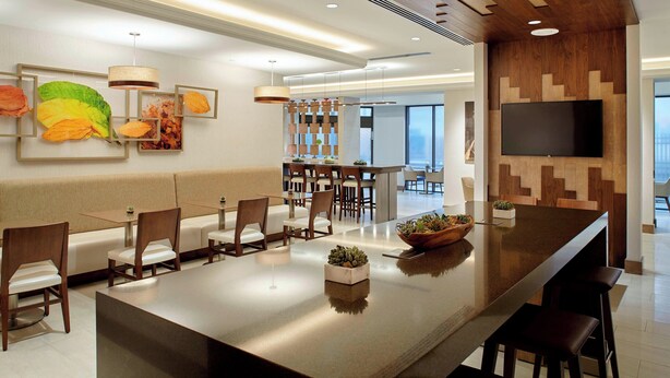A modern hotel lobby with a long bar, seating area, and a TV mounted on the wall.