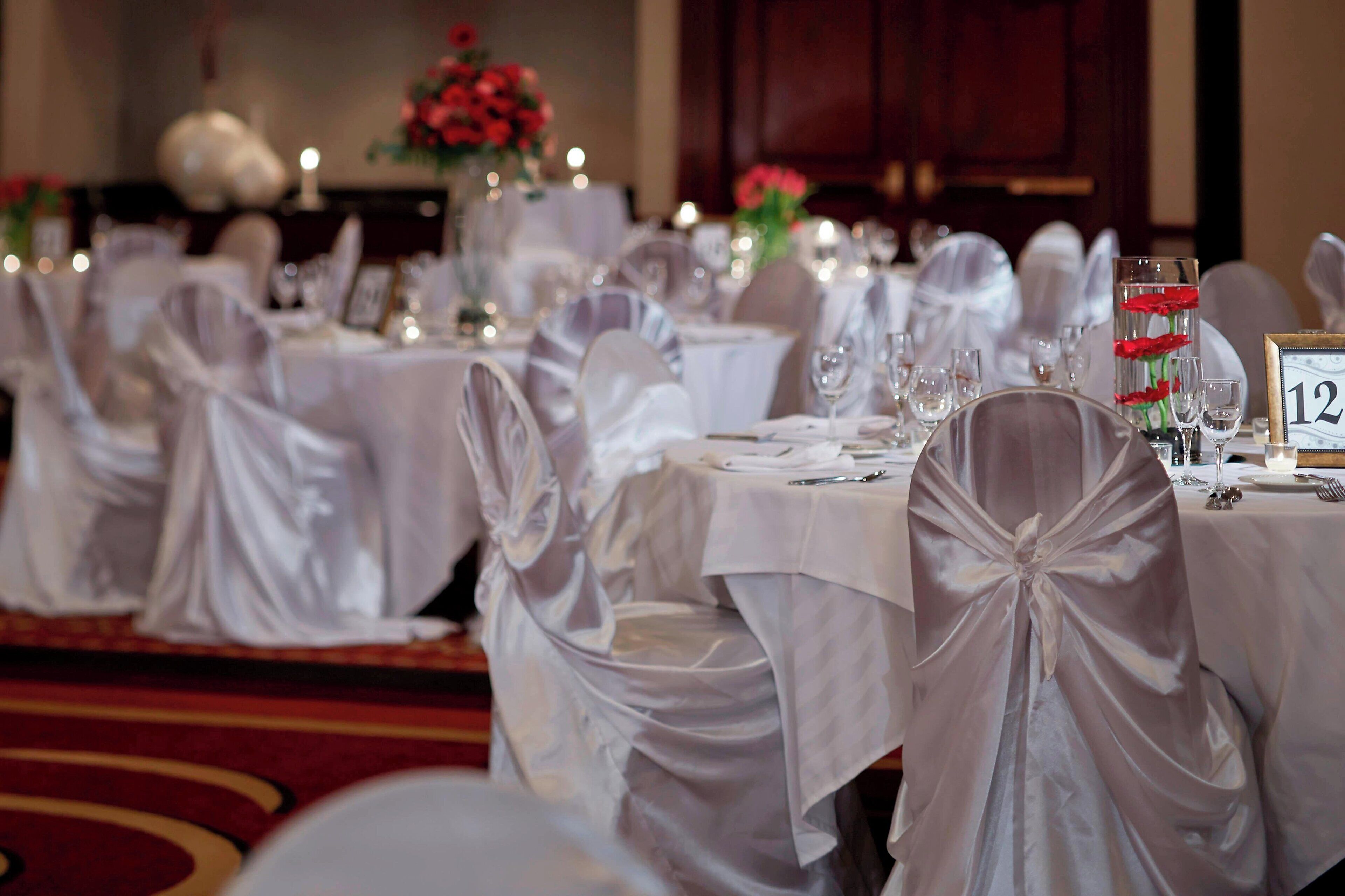 A banquet hall with round tables covered in white tablecloths, each with a number sign. Chairs are covered with white fabric and tied with bows.