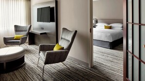 A hotel room with a modern design, featuring a large bed, two armchairs with cushions, a flat-screen TV mounted on the wall, and a carpeted floor.