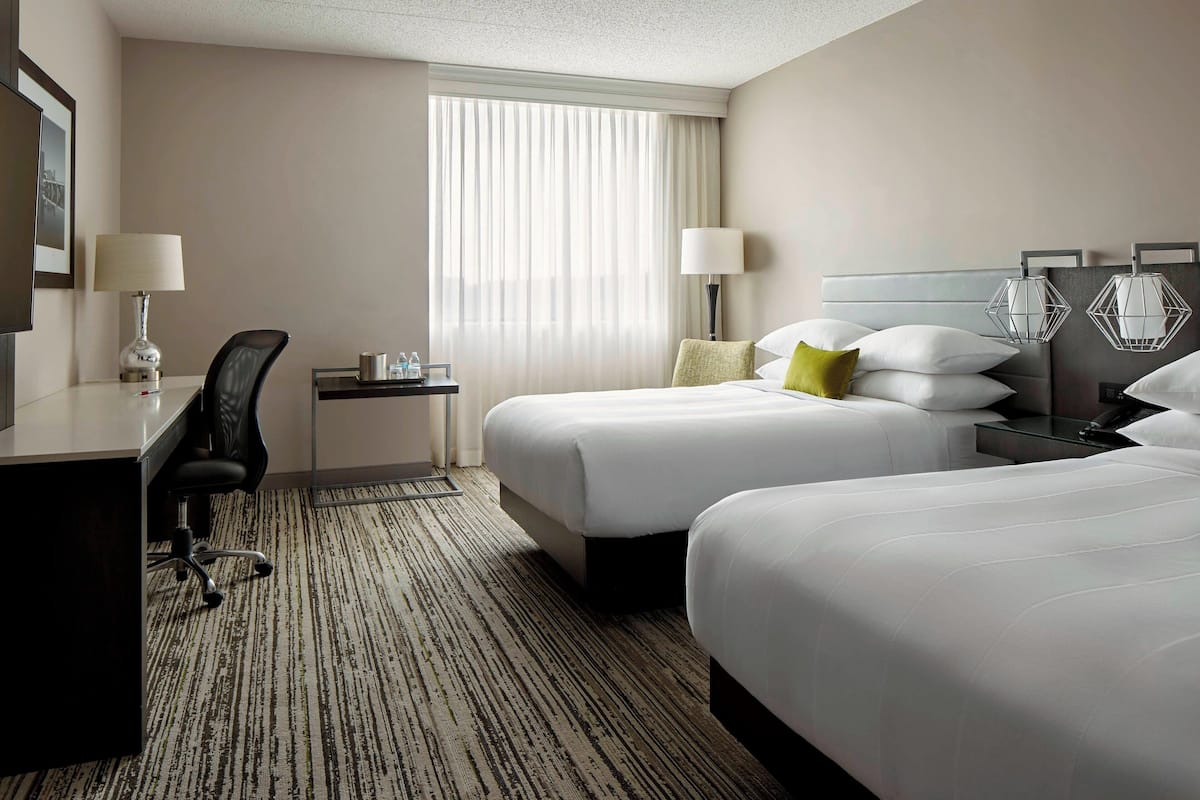 Une chambre d’hôtel avec deux lits, un bureau, une chaise et une fenêtre avec des rideaux légers.