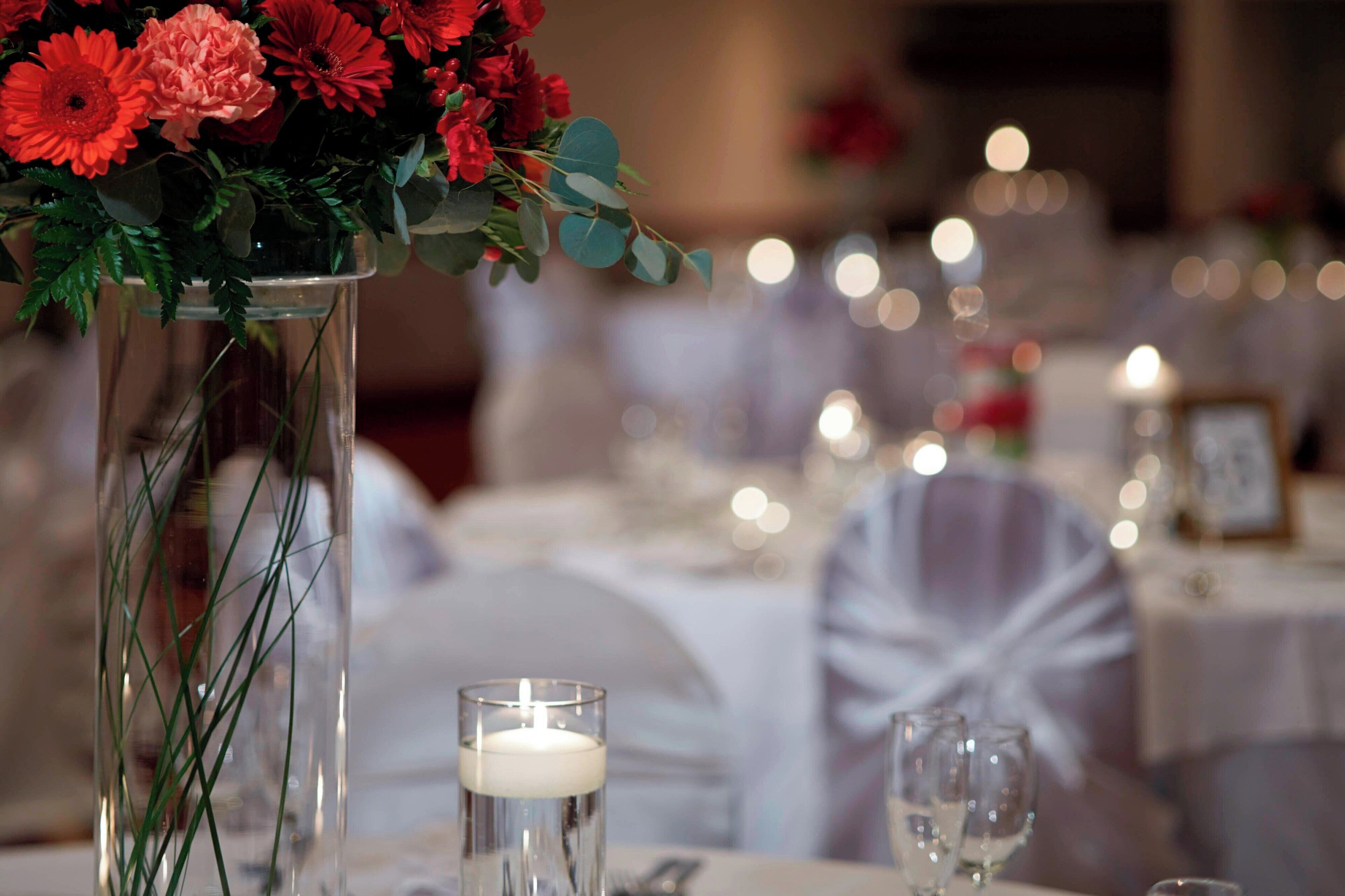 Une table dressée avec un grand vase en verre contenant des fleurs rouges et des feuilles vertes, une bougie allumée et de la verrerie transparente.