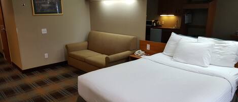 A hotel room with a bed, a sofa, a small table with a phone, a painting on the wall, and a kitchenette area.