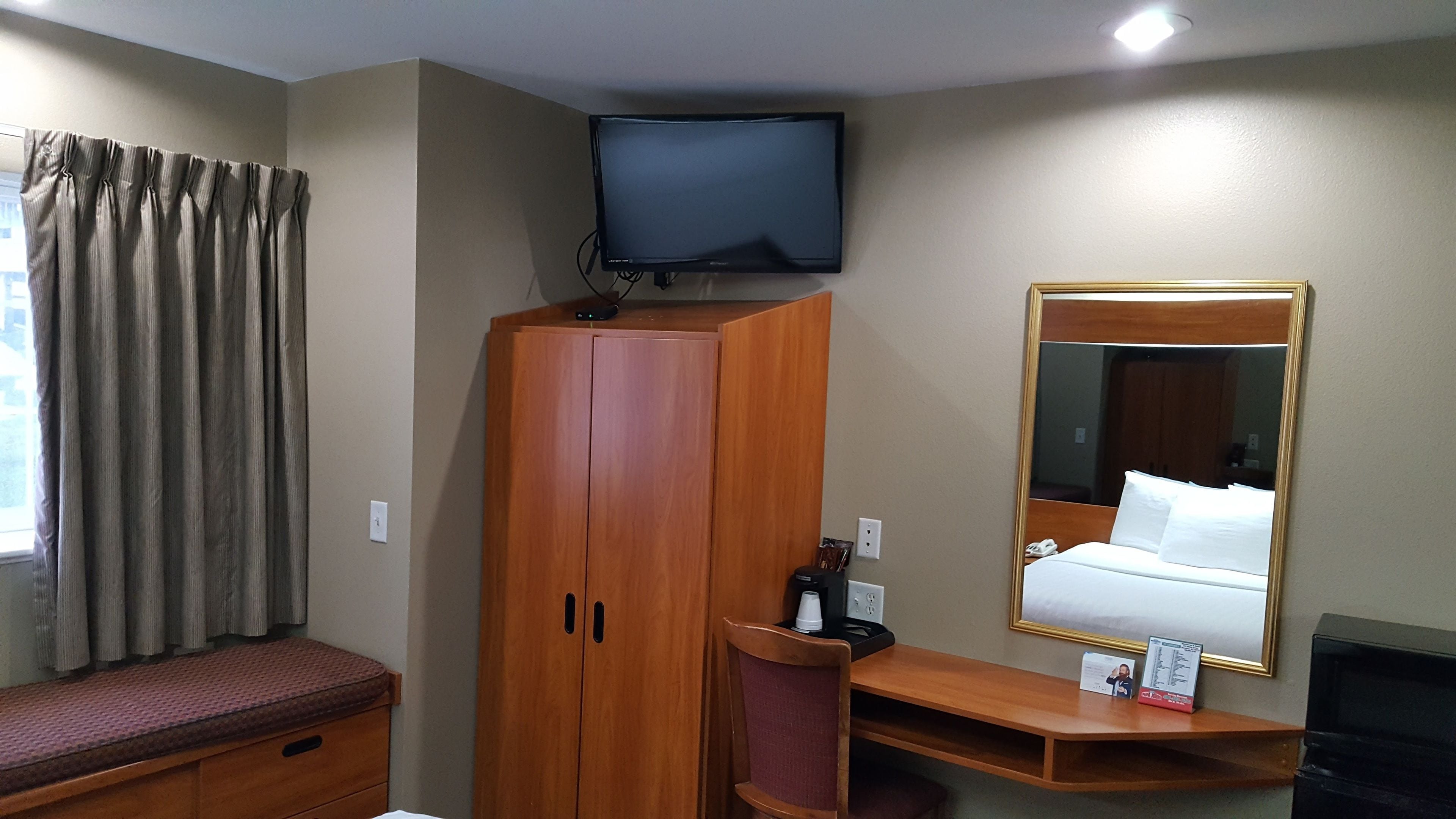 A hotel room with a wooden wardrobe, a television mounted above it, a bed with a headboard, a bedside table with a phone and a book, and a window with curtains.