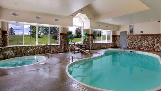 An indoor swimming pool with a hot tub, stone walls, and large windows offering a view of the outdoors. - Microtel Inn & Suites by Wyndham Bozeman (Bozeman)
