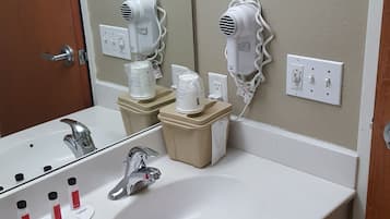 A bathroom with a white sink, two hair dryers mounted on the wall, a mirror, and a trash bin.
