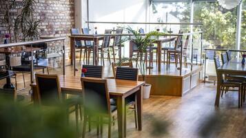 Un café espacioso con mesas y sillas de madera, amplios ventanales que ofrecen vistas al verdor exterior, y plantas en macetas que complementan la decoración.