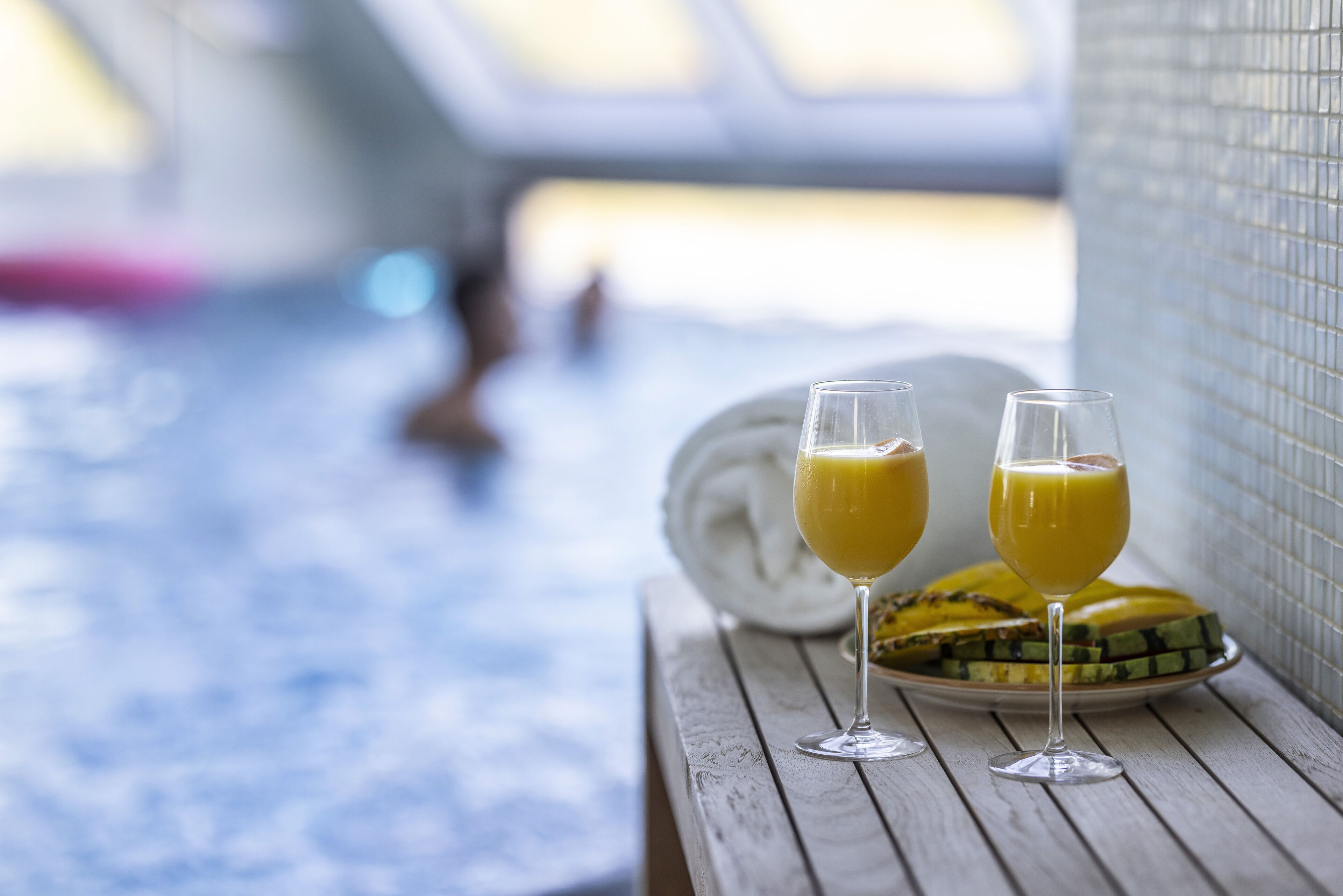 Una mesa de madera con dos vasos de jugo de naranja y un plato de fruta cortada, probablemente sandía, cerca de una zona de piscina con un muro de azulejos y una persona al fondo.
