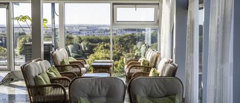 A sunlit indoor seating area with wicker chairs and tables, large windows offering a view of the cityscape.