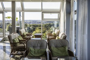 A sunlit indoor seating area with wicker chairs and tables, large windows offering a view of the cityscape.