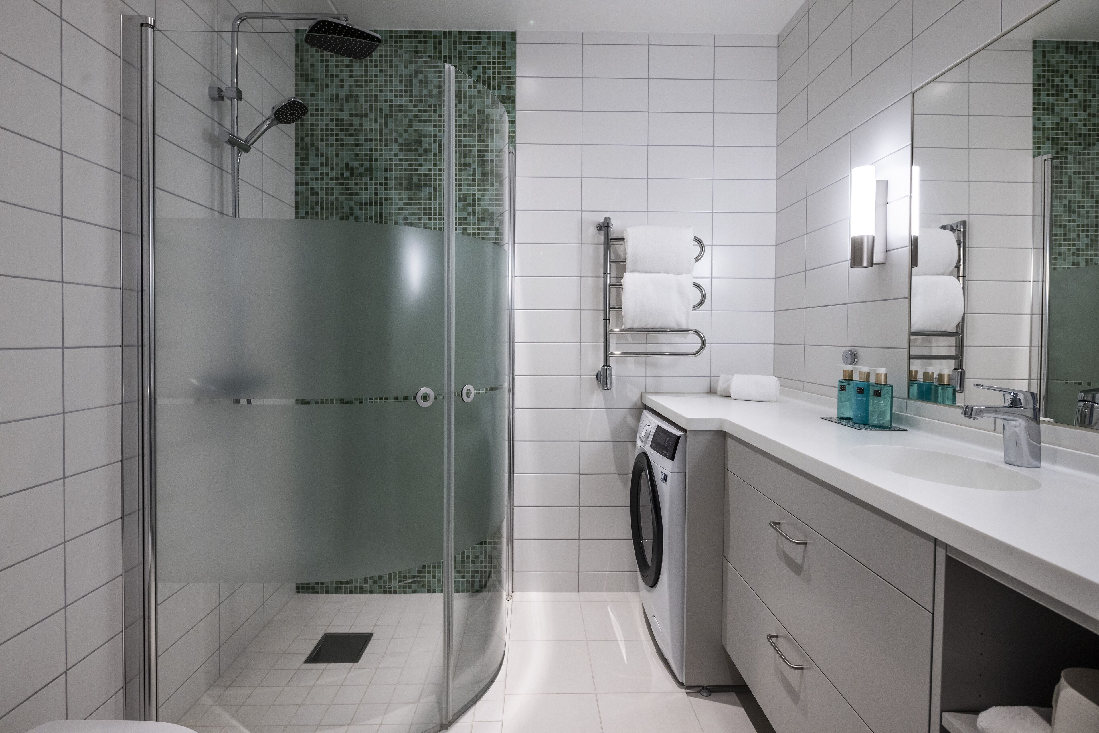 A modern bathroom with a glass shower enclosure, a built-in washing machine, and a large mirror.