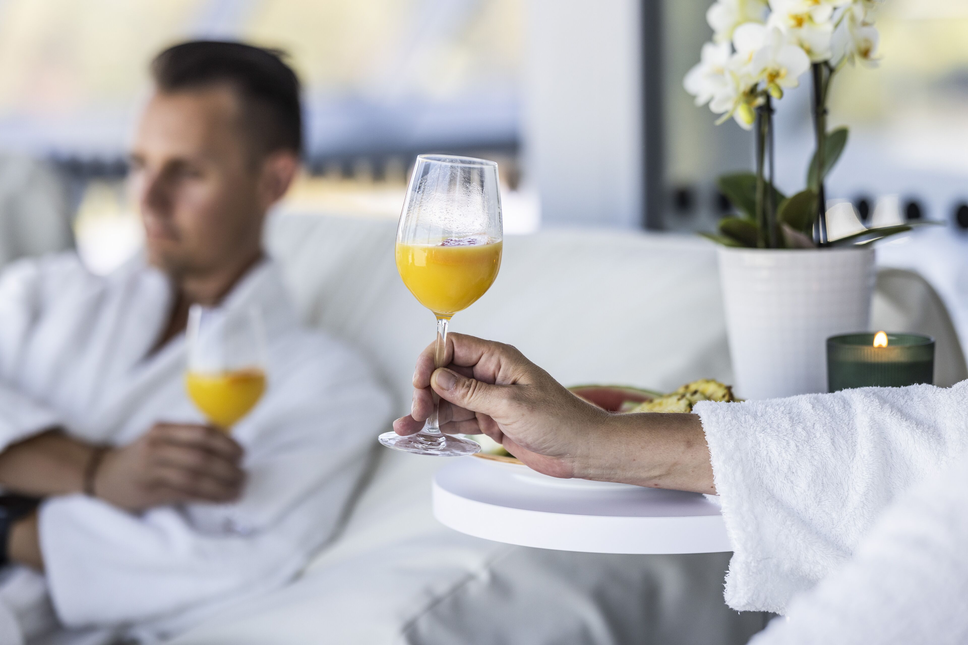 A person holding a glass of orange juice, another person in the background holding a glass, a white towel, and a candle on a table.