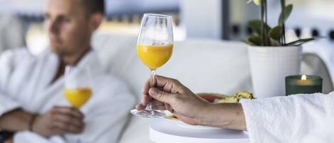 Una persona sosteniendo un vaso de jugo de naranja, otra persona al fondo con un vaso, una toalla blanca y una vela sobre una mesa.