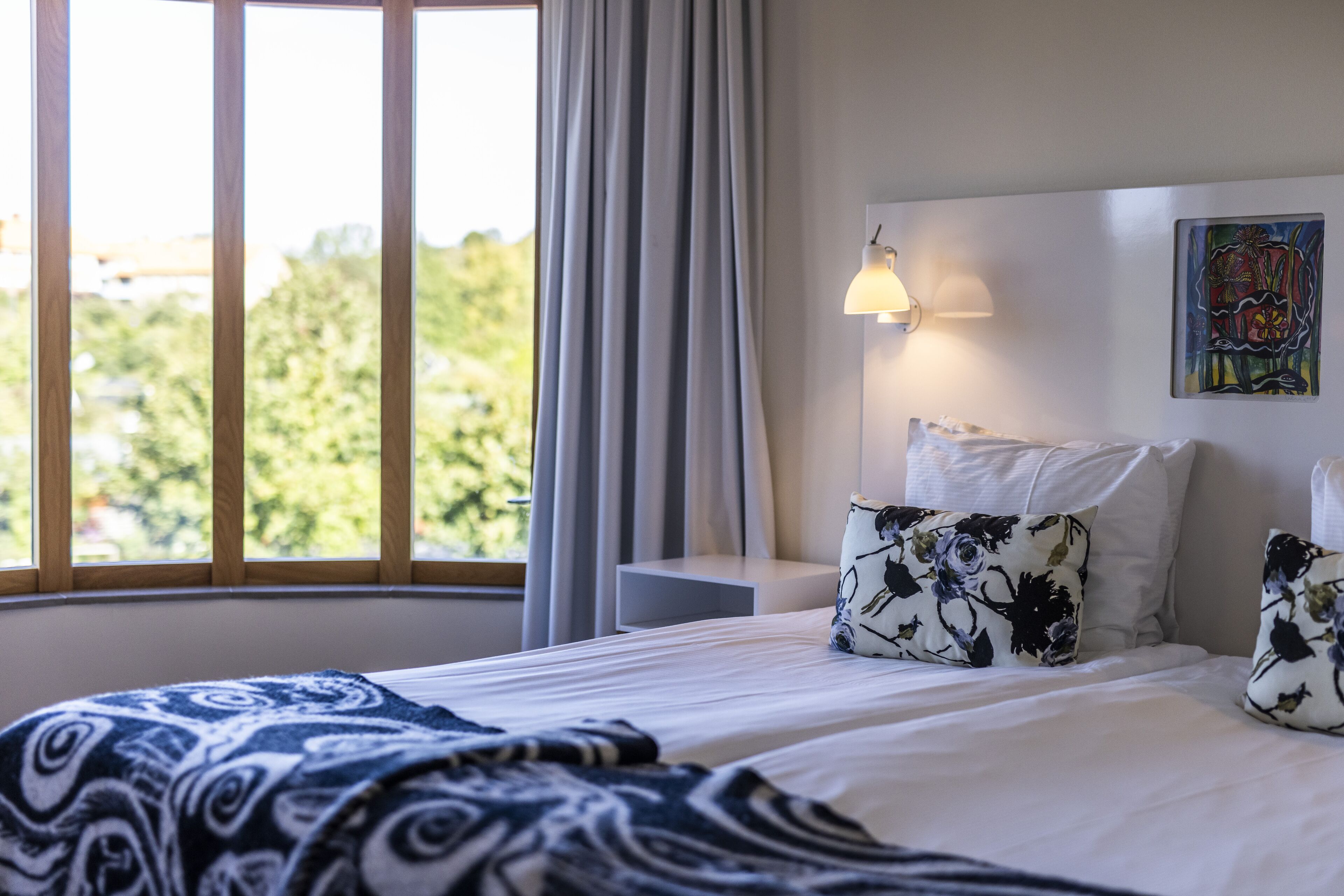 Une chambre d’hôtel avec une grande fenêtre, un lit recouvert de draps blancs et agrémenté d’oreillers à motifs floraux, une table de chevet avec une lampe et un tableau accroché au mur.