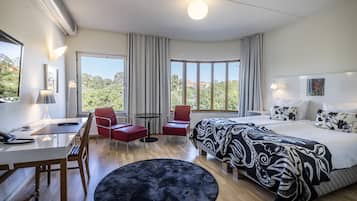 Hotelkamer met een bed, een bureau, twee rode stoelen, een klein tafeltje en een groot raam met uitzicht op groen.