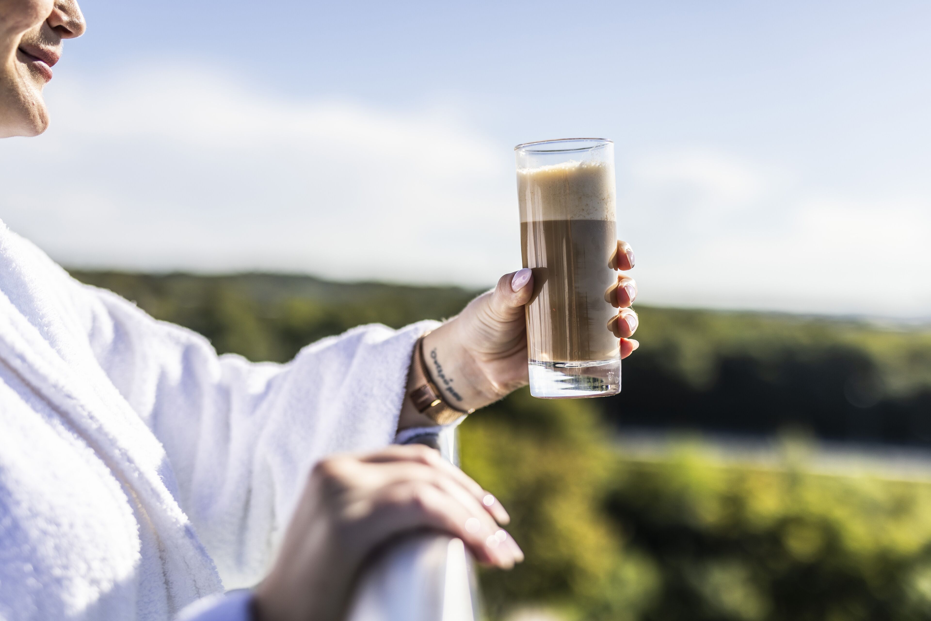 A person holding a tall glass with a beverage, wearing a white robe, and standing by a railing with a natural landscape in the background.