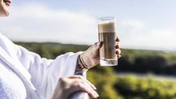 A person holding a tall glass with a beverage, wearing a white robe, and standing by a railing with a natural landscape in the background.