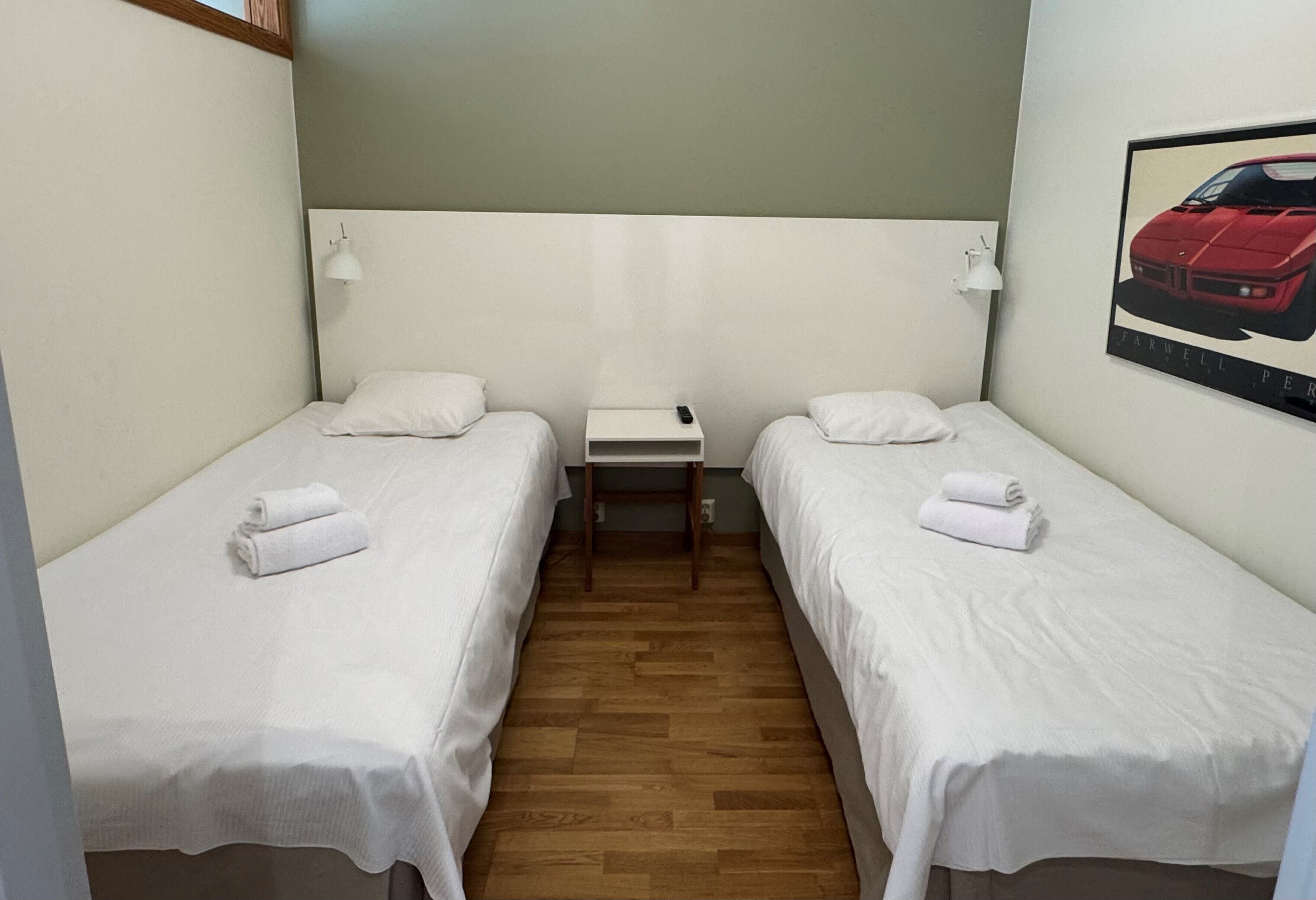 Una habitación pequeña y limpia con dos camas individuales, una mesita y una lámpara de pared.
