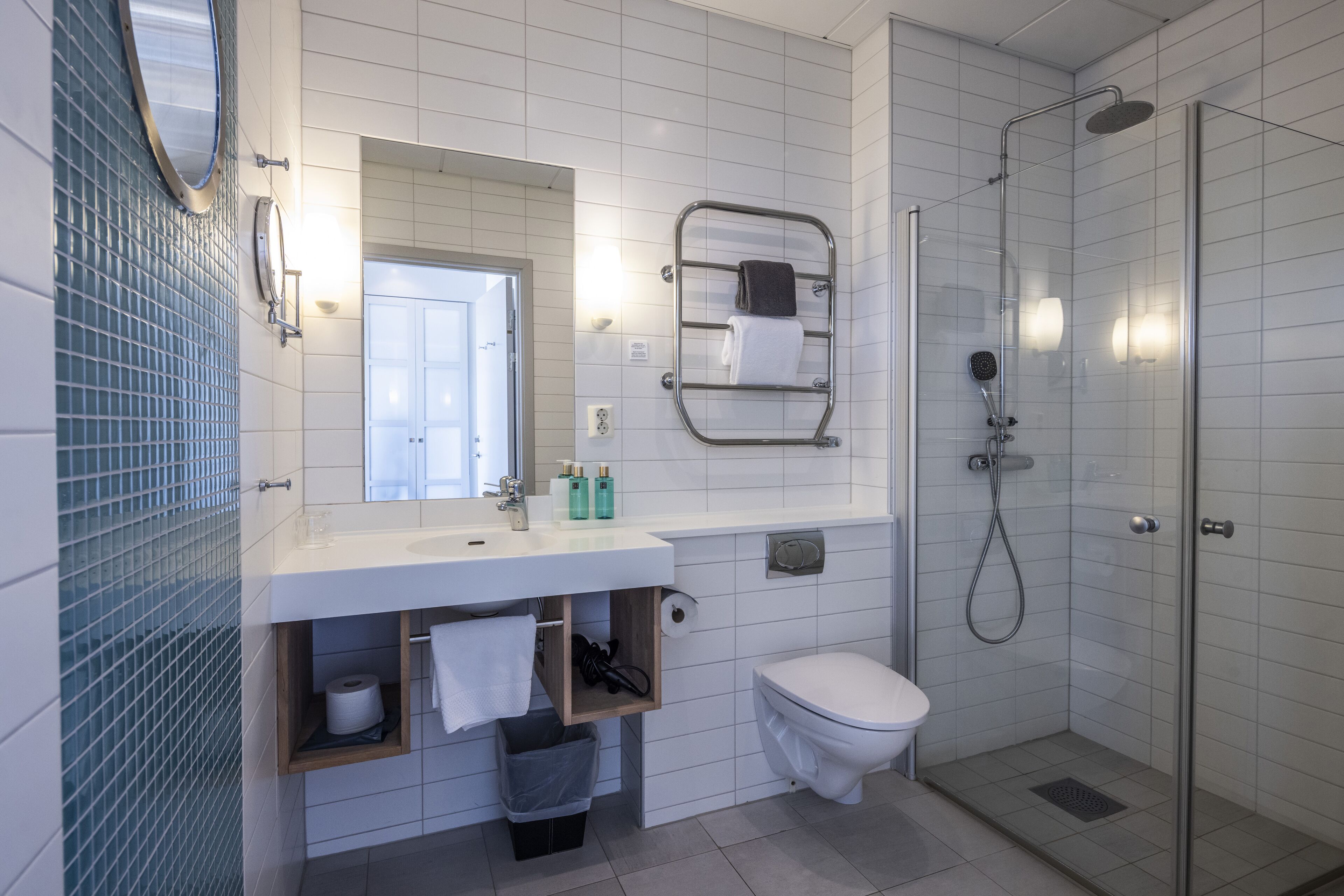 Un baño moderno con lavamanos, espejo y retrete blancos, cabina de ducha con mampara de vidrio y un perchero para toallas.