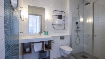 Un baño moderno con lavamanos, espejo y retrete blancos, cabina de ducha con mampara de vidrio y un perchero para toallas.