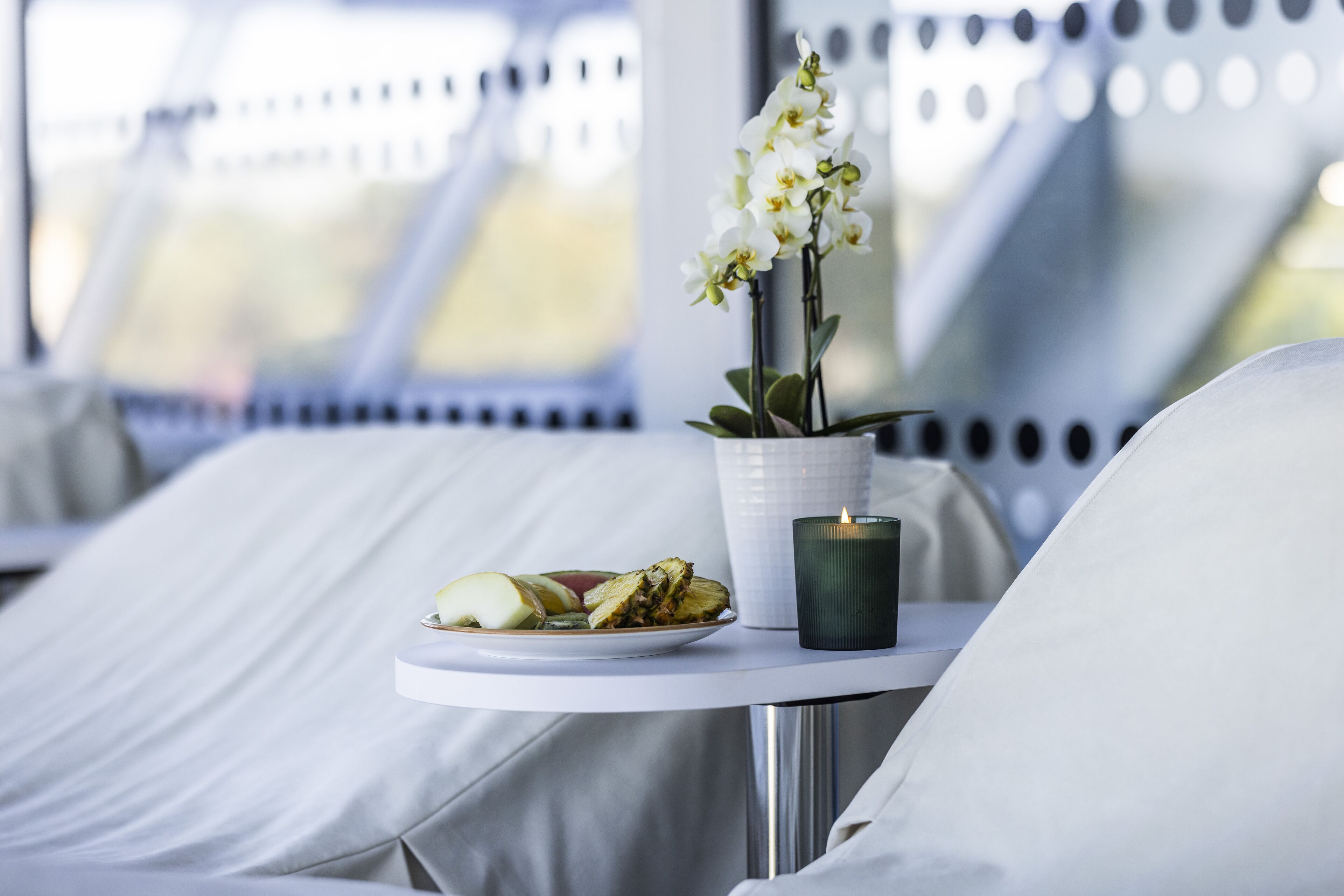 Una cama con sábanas blancas, una mesita con una vela, un plato de comida y una orquídea en maceta.